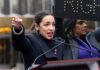 AOC Drops “Genocide” Bomb in NAZI Birthplace A woman passionately speaking at a political rally with a sign in the background