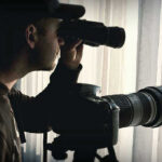 Person looking through binoculars beside a camera.