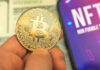 Person holding Bitcoin coin, NFT text on screen.