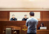 Plane Hijacker Walks Free — Judge Blamed Man stands in courtroom before judges and stenographer.