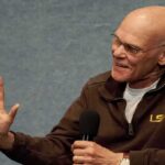 James Carville speaking into a handheld microphone, gesturing with his left hand