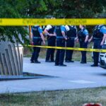 Columbia Officer Killed in Park Shooting police officers stand behind caution tape at a park scene