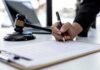Judge Blocks Trump’s Firing of U.S. Attorney Hand signing legal document beside gavel and laptop