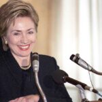 Hillary Clinton smiling at a podium with multiple microphones