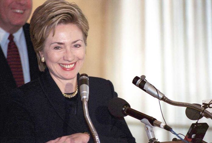 Hillary Clinton smiling at a podium with multiple microphones